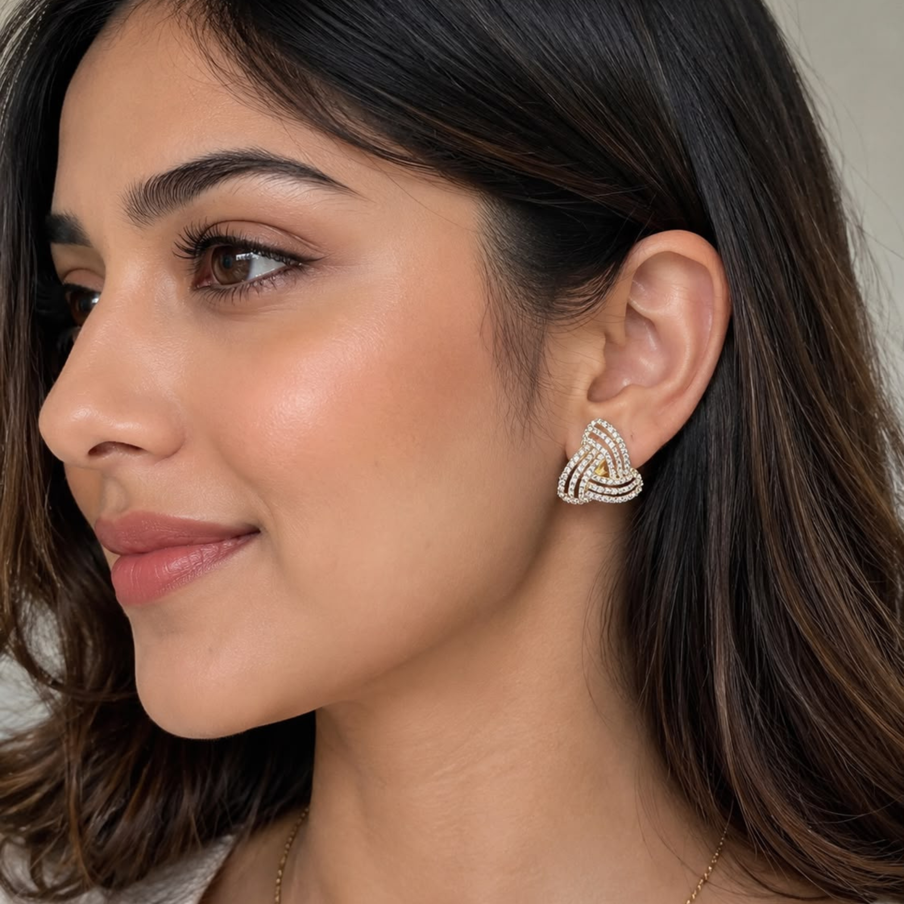 A close-up of a woman wearing a small triquetra-style stud earring with a gold center and silver outer loops, embellished with small clear stones.